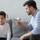 A man sits on a couch and sternly points his finger at his son who looks away.